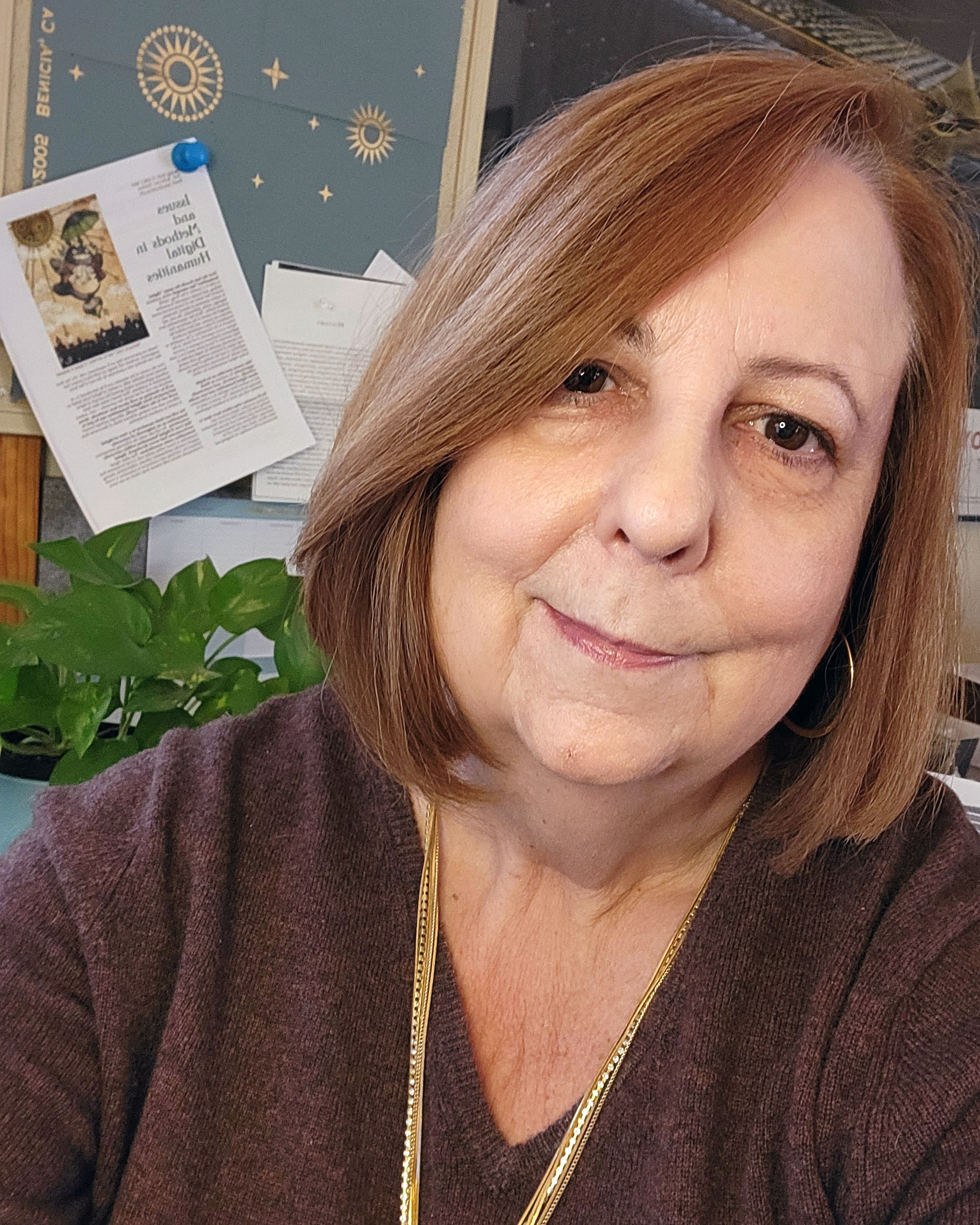 Katherine Pandora has shoulder-length brown hair and is smiling softly. She wears a brown top and gold necklace and sits in an office setting. Behind her is a green plant and bulletin board displayhing papers.
